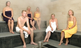 Sechs Gäste sitzen auf der Steinbank, Wasserdampf steigt auf - SinnBad im Classic-Saunabereich Sechs Gäste sitzen auf der Steinbank, Wasserdampf steigt auf - SinnBad im Classic-Saunabereich
